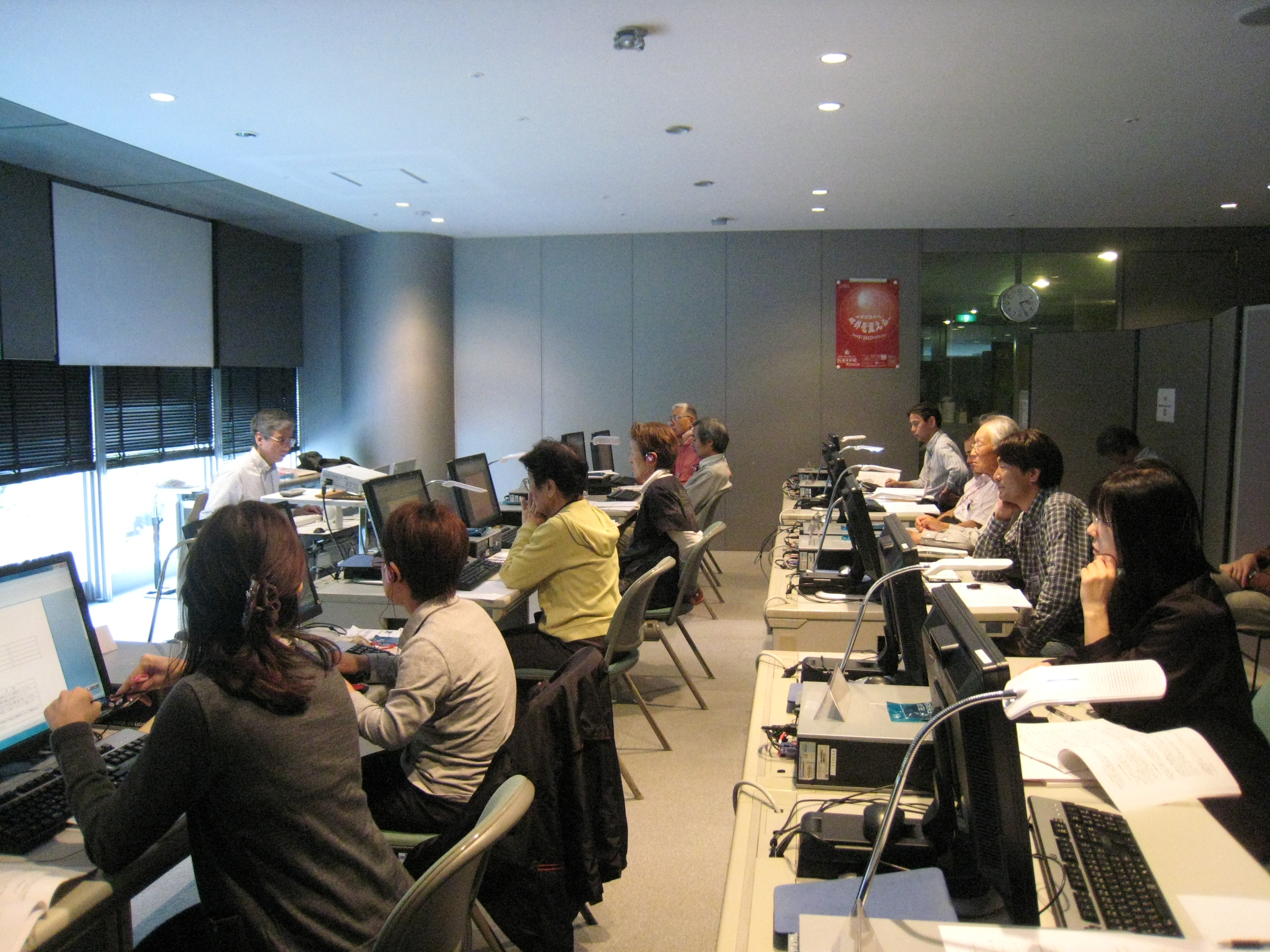 写真注:東京会場は」11名の受講者と講師の姿。そして講師の斜め後ろにSkypeでの共有画像を映し出すスクリーンがあります。塩尻会場は東京から中継されてきた画像を映し出すスクリーンがあり、5名の受講者がそれを注視しています。注終わり